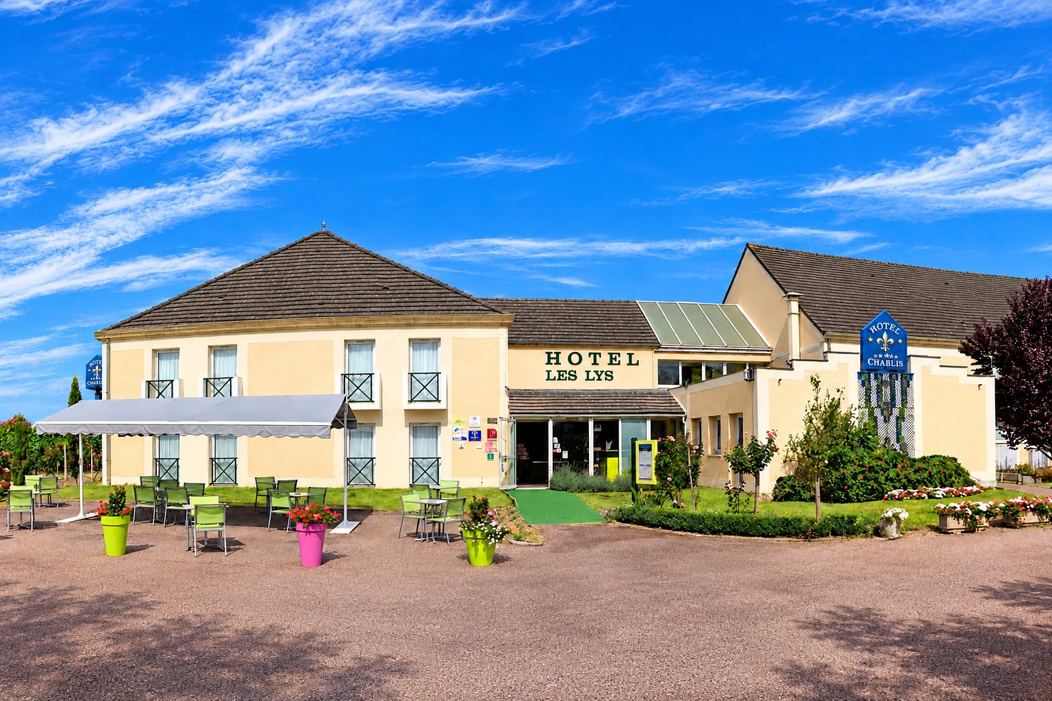 Façade beige de l’hôtel Les Lys avec son enseigne, une terrasse équipée de tables et chaises vertes et des massifs de fleurs au premier plan.
