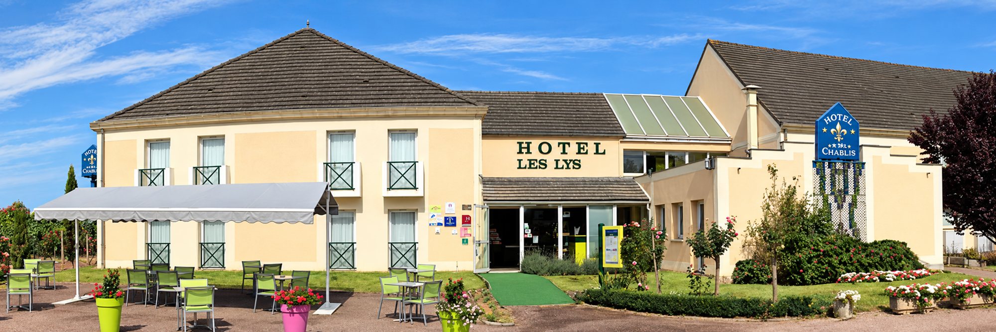 Façade beige de l’hôtel Les Lys avec son enseigne, une terrasse équipée de tables et chaises vertes et des massifs de fleurs au premier plan.