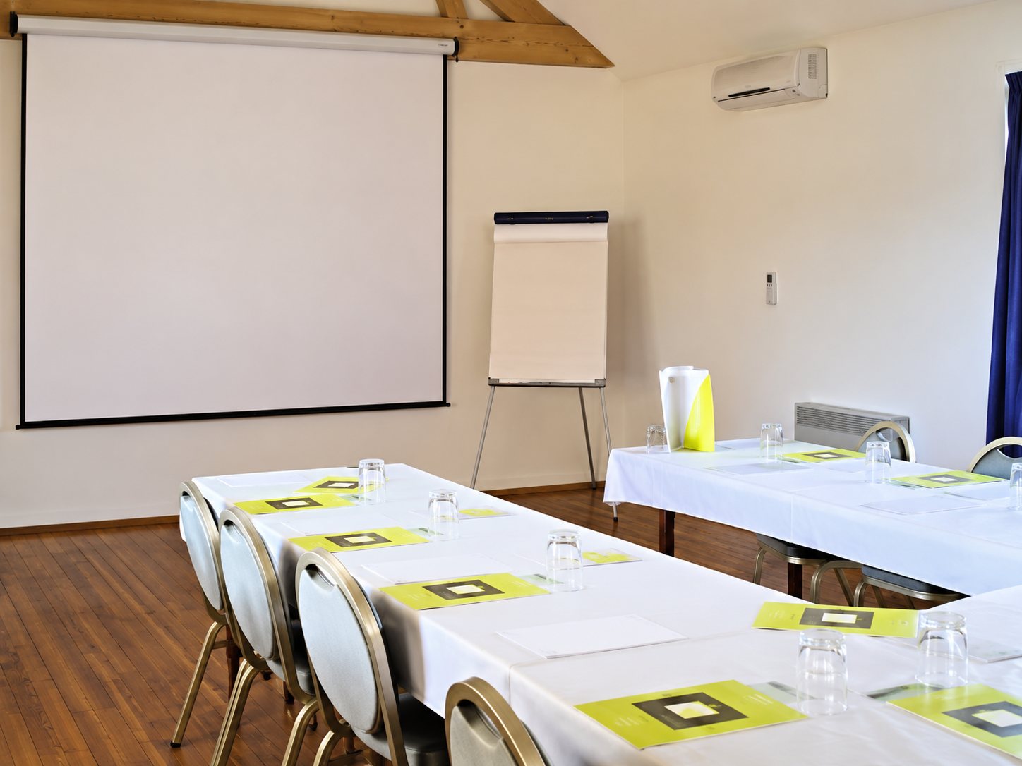 Une salle de réunion lumineuse avec de longues tables garnies de cartons verts, un grand écran blanc et un paperboard au fond.