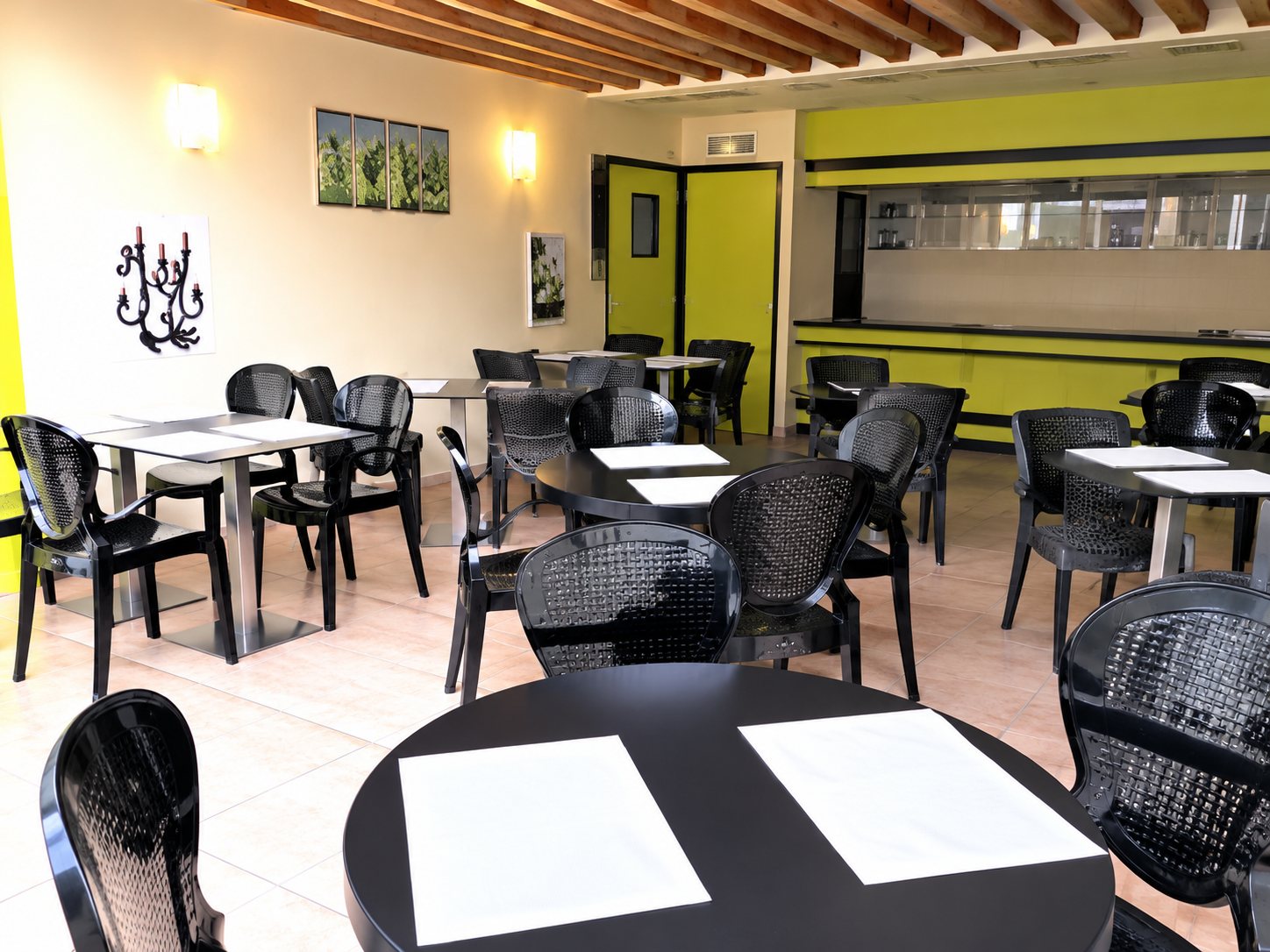 Intérieur lumineux d’un restaurant avec plusieurs tables carrées et des chaises noires, des sets de table blancs posés au premier plan.